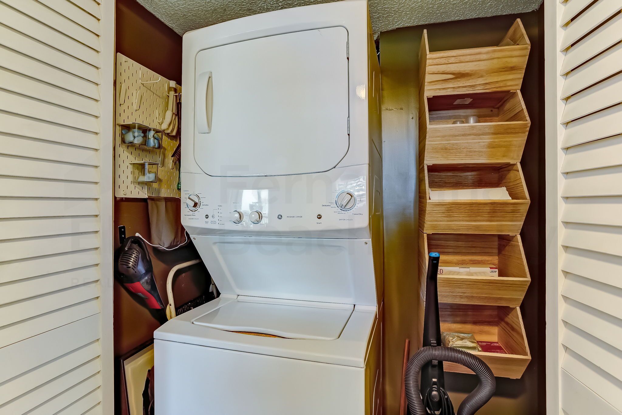 A compact laundry room with a front-loading washer and dryer, a drying rack, and a wooden storage unit with multiple shelves.