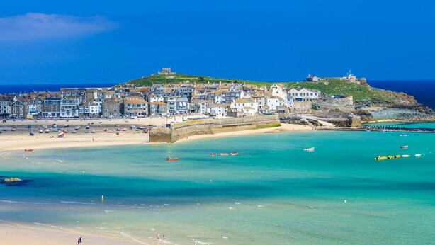 A coastal town with a beach, clear blue waters, and a hilltop building. - 15 Tower Meadows (Penzance)