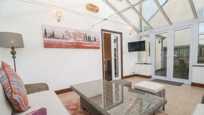 A modern living room with a glass table, wicker furniture, a flat-screen TV, and a painting on the wall. - 15 Tower Meadows (Penzance)