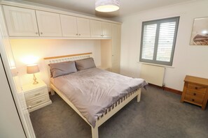 A bedroom with a bed, nightstand, lamp, window, and built-in wardrobe. - 15 Tower Meadows (Penzance)