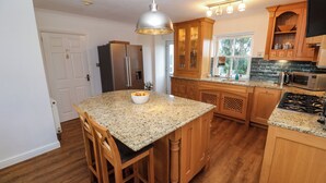 A kitchen with wooden cabinets, granite countertops, stainless steel appliances, and a large island with bar stools. - 15 Tower Meadows (Penzance)