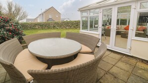 A round wicker table with cushioned chairs in a covered outdoor area with a stone wall and a house in the background. - 15 Tower Meadows (Penzance)