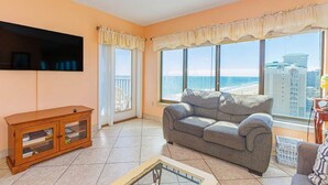 Uma sala de estar com sofá, televisão na parede, armário de madeira e grandes janelas com vista para o mar e edifícios.