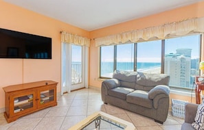 A living room with a sofa, a TV mounted on the wall, a wooden cabinet, and large windows with a view of the ocean and buildings.