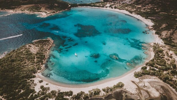 Vista aérea de una zona costera con playa de arena, aguas turquesas cristalinas y algunas embarcaciones.