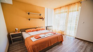 Quarto moderno com piso de madeira, cama com roupa de cama laranja, mesa de cabeceira com abajur e janela com cortinas translúcidas.