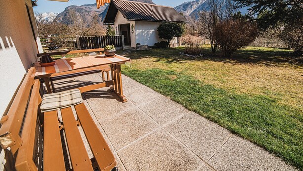 Um pátio com bancos de madeira e uma mesa, uma casa pequena com telhado de metal, e vista para as montanhas ao fundo.