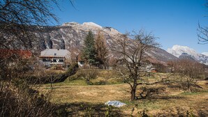 Uma casa com telhado inclinado, cercada por um gramado com árvores e montanhas ao fundo.