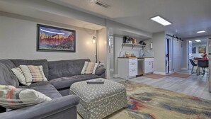 A modern living room with a grey sofa, ottoman, and colorful rug. There is a kitchen area with white cabinets and a dining table. - On-Site Trails! Hot Tub Hideaway in Zion Area (Hurricane)
