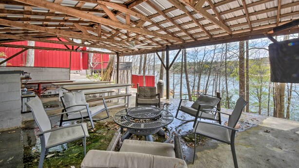 A covered outdoor seating area with metal chairs and a fire pit, overlooking a body of water and a red building in the background. - Lakefront Lavonia Cottage w/ 2-Story Dock! (Lavonia)