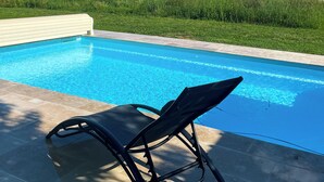 En swimmingpool med en sort liggestol, omgivet af græs og træer.