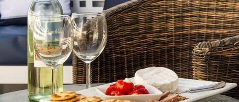 A wicker chair, a glass table, a bottle of white wine, a glass, and a plate with cheese, crackers, and strawberries.
