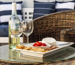 Een rieten stoel, een glazen tafel, een fles witte wijn, een glas en een bord met kaas, crackers en aardbeien.