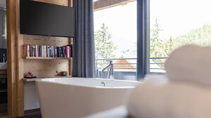 A modern bathroom with a freestanding bathtub, wooden shelves with books, and a view of a forested area through a large window. - Superior Chalet for 10 people with sauna (Turrach)