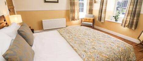 A well-lit bedroom with a large bed, a bedside table, a chair, a window with curtains, and a radiator.