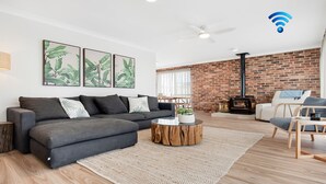 A modern living room with a grey sofa, a round wooden coffee table, a brick wall, a white ceiling fan, and a wood-burning stove. - Blondie's – beach at the end of the street (Culburra Beach)
