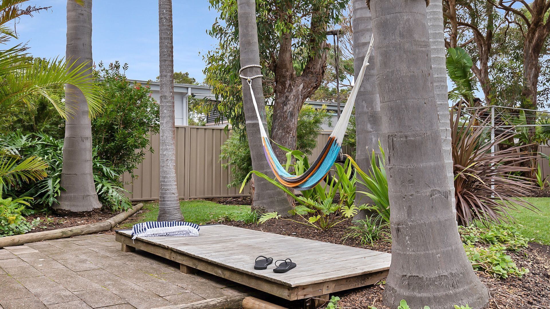 A hammock between trees, a wooden deck with a blanket and flip-flops, and a wooden fence in a garden.
