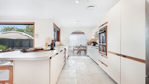A modern kitchen with a central island, a dining area with a table and chairs, and a view of the outdoors. - Blondie's – beach at the end of the street (Culburra Beach)