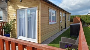 Une cabane en rondins dotée de grandes portes-fenêtres coulissantes, d’une antenne parabolique et d’un petit espace jardin aménagé avec une chaise en osier et des plantes en pot.