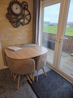 A dining area with a round wooden table, two cushioned chairs, a decorative wall clock, and a view of a grassy outdoor area through a glass door.