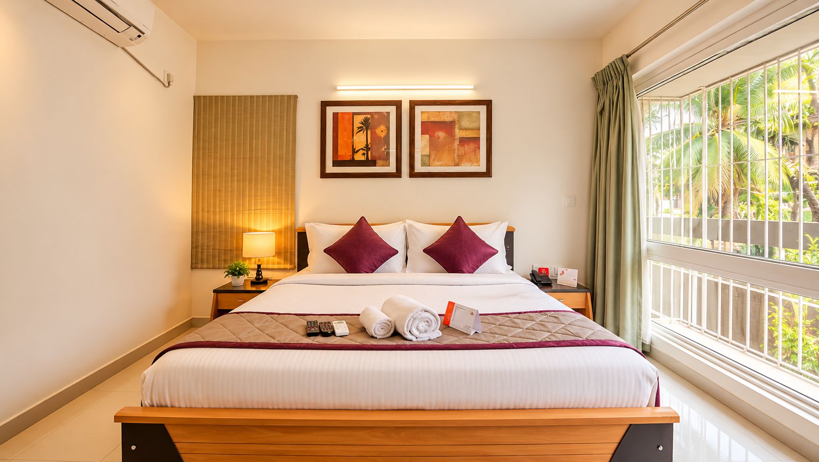 A hotel room with a bed, two bedside tables, a window with a view of palm trees, and two framed pictures on the wall.