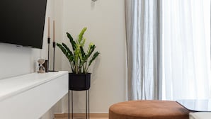 Un soggiorno moderno con una televisione a schermo piatto montata a parete, una pianta in vaso, un poggiapiedi rotondo e un tavolino consolle bianco.