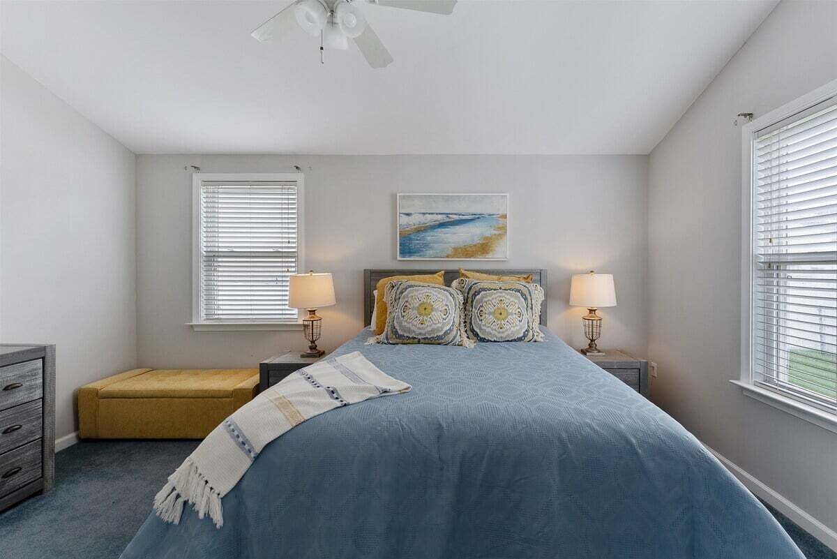 Quarto com cama, duas luminárias de cabeceira, ventoinha no teto, janela com persianas, um pequeno banco e um quadro na parede.