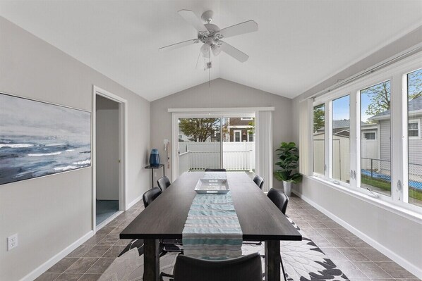 A dining area with a long table, chairs, a ceiling fan, and a view through large windows.