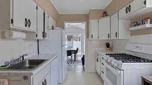 Een keuken met witte kasten, een roestvrijstalen spoelbak, een fornuis, en een eethoek zichtbaar door een open deur.