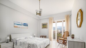 Un dormitorio con cama, mesitas de noche, una silla, escritorio, espejo y balcón con cortinas.