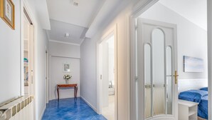 Un pasillo con suelo de baldosas azules, paredes blancas y una puerta que da a un dormitorio con cama y manta azul.