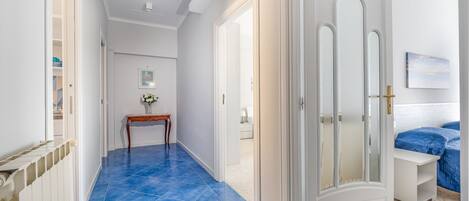A hallway with blue-tiled flooring, white walls, and a door leading to a bedroom with a bed and a blue blanket.
