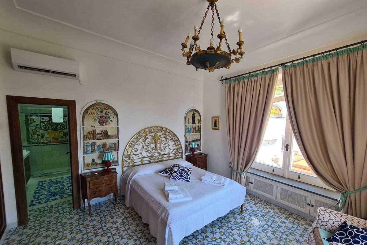 Une chambre à coucher avec un lit, un lustre, une fenêtre avec des rideaux, un miroir et un sol à motifs.