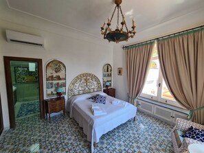 A bedroom with a bed, a chandelier, a window with curtains, a mirror, and a patterned floor.