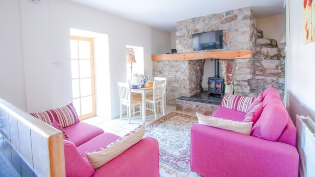 A living room with a stone fireplace, a television mounted on the wall, and a dining area with a table and chairs. - Berry Hall - Porty Eynon (Swansea)