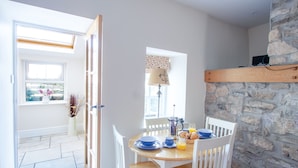 A dining area with a stone wall, a wooden table, white chairs, and a window with a view of greenery. - Berry Hall - Porty Eynon (Swansea)