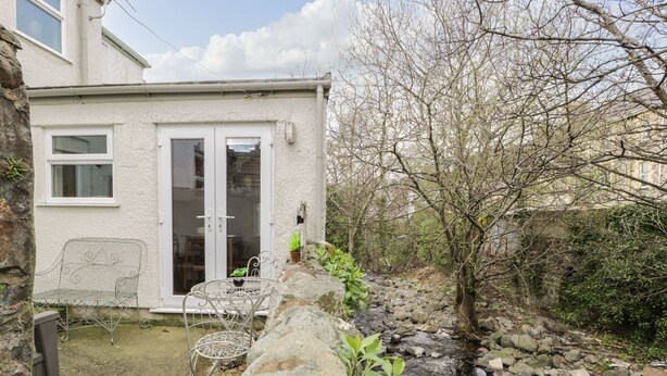Un mur de pierre menant à une terrasse avec une table et des chaises, un bâtiment blanc doté d’une porte en verre et un ruisseau rocheux.