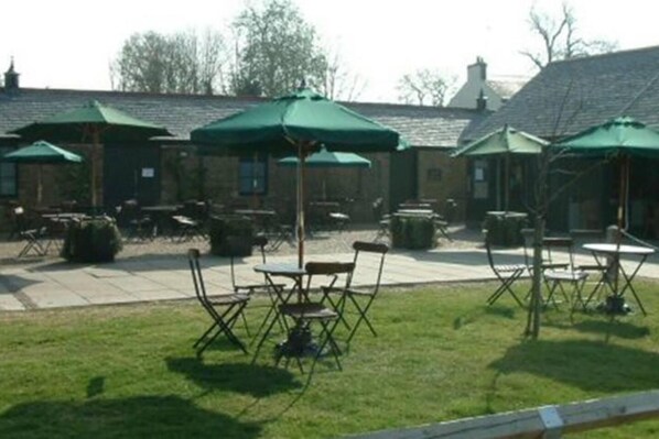 An outdoor seating area with tables and chairs under green umbrellas, a grassy lawn, and a building with a brick facade in the background.