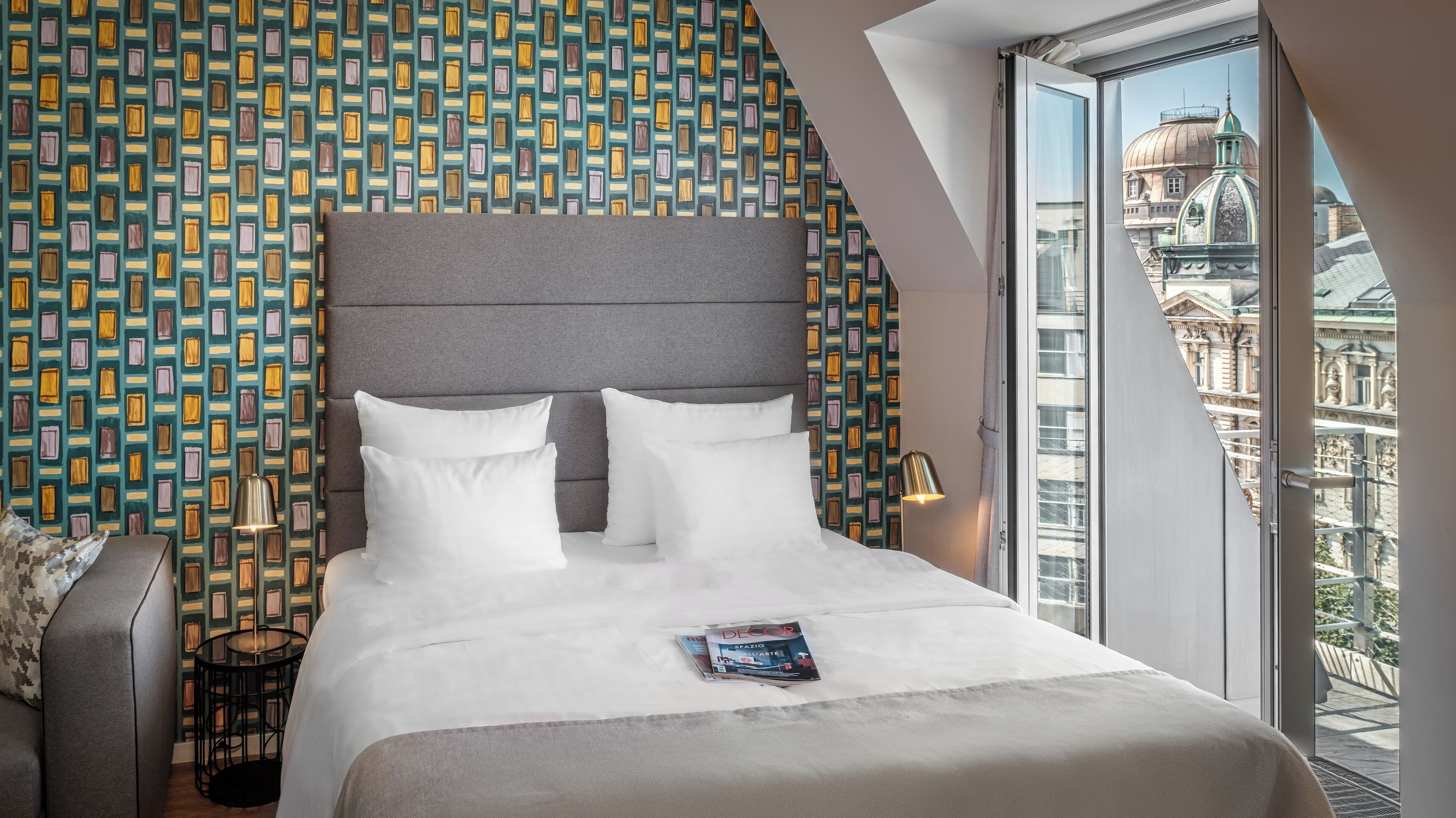 Een hotelkamer met een groot bed, een behang met patroon, uitzicht op een stadsbeeld en een balkon.