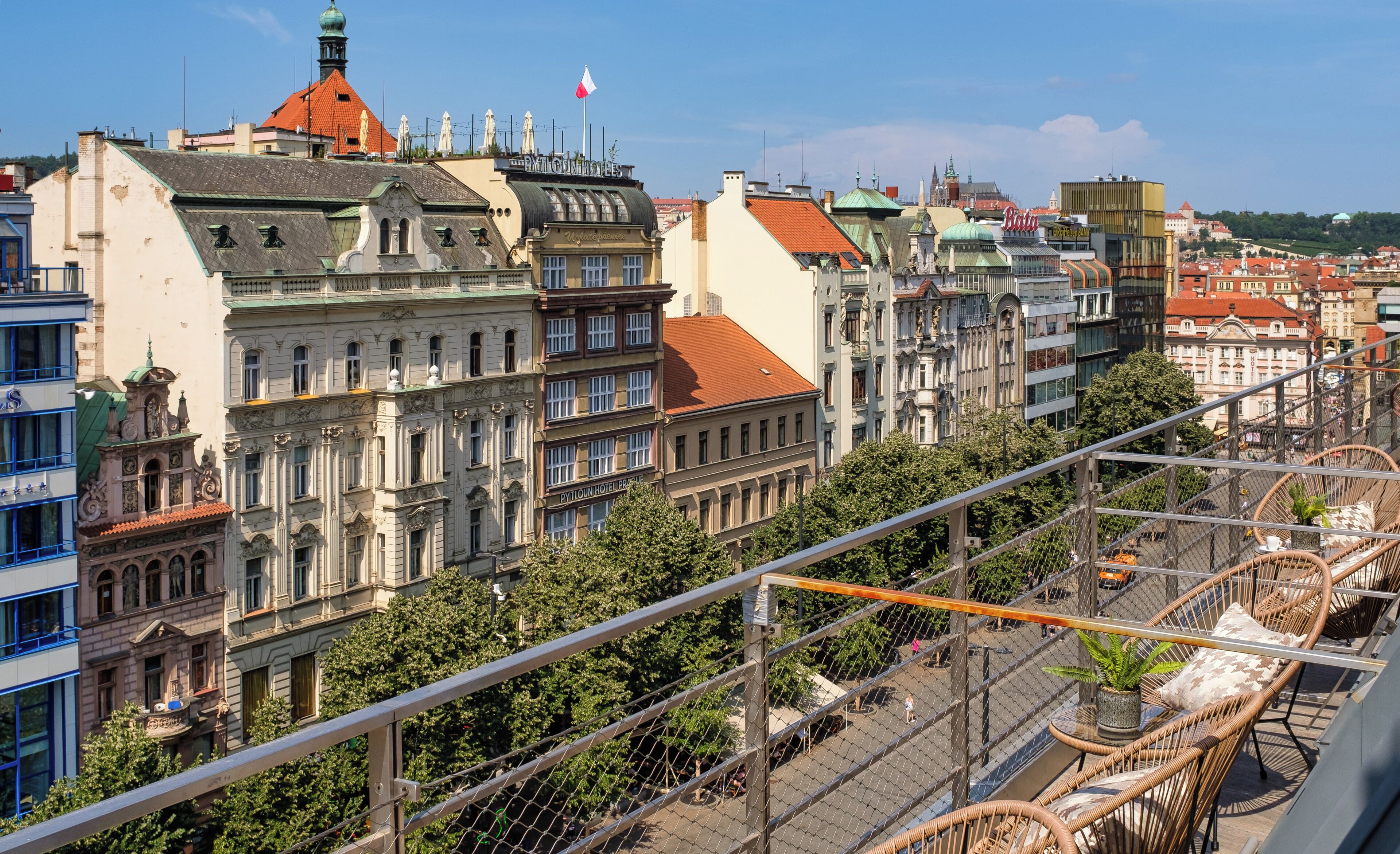 A view of a cityscape with historic buildings, including a hotel with a red roof and a sign that reads 'Hotel Prague.'