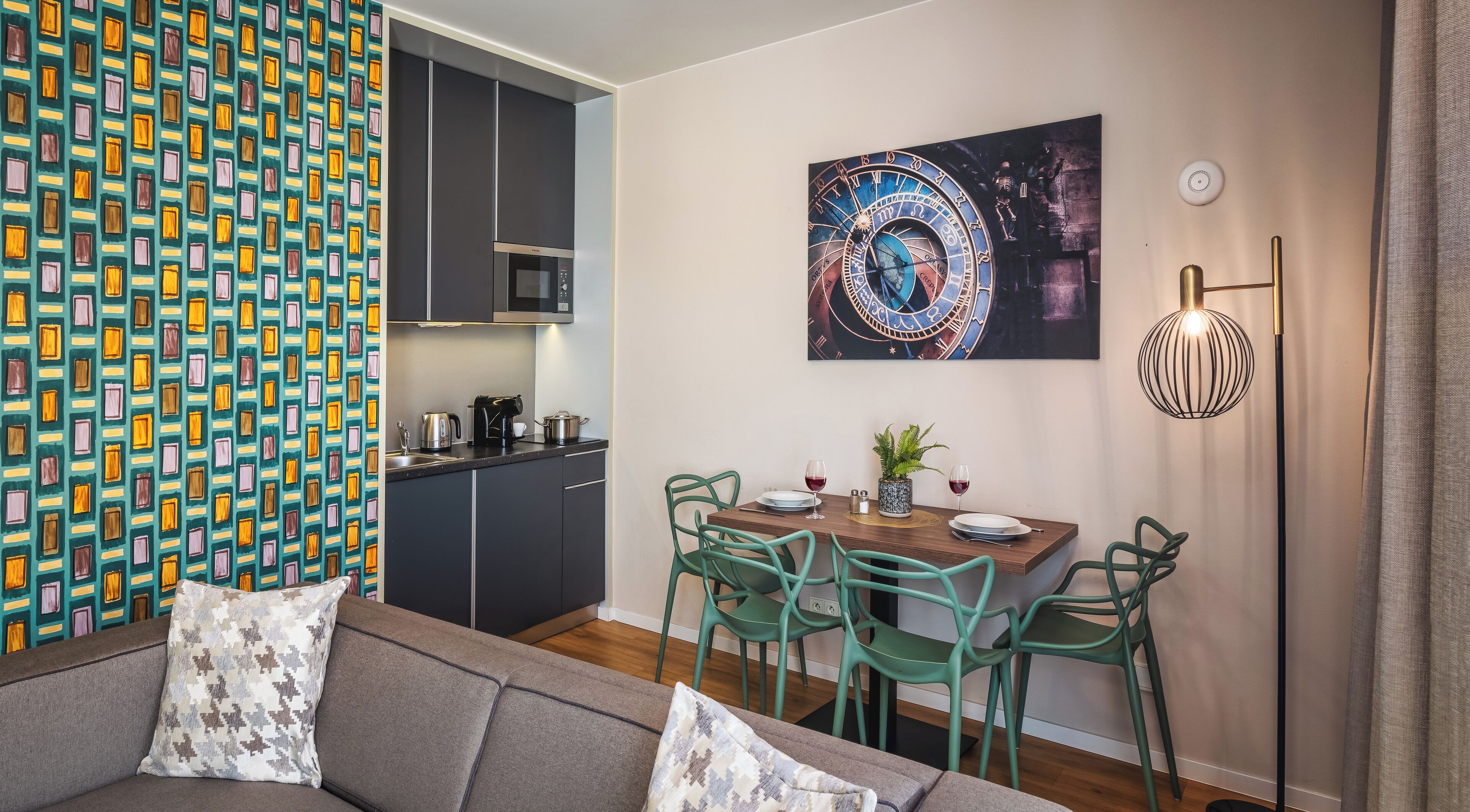 Un salon moderne avec un canapé et des fauteuils verts, une table à manger avec des assiettes et des verres, une cuisine avec des meubles sombres et une grande horloge murale.