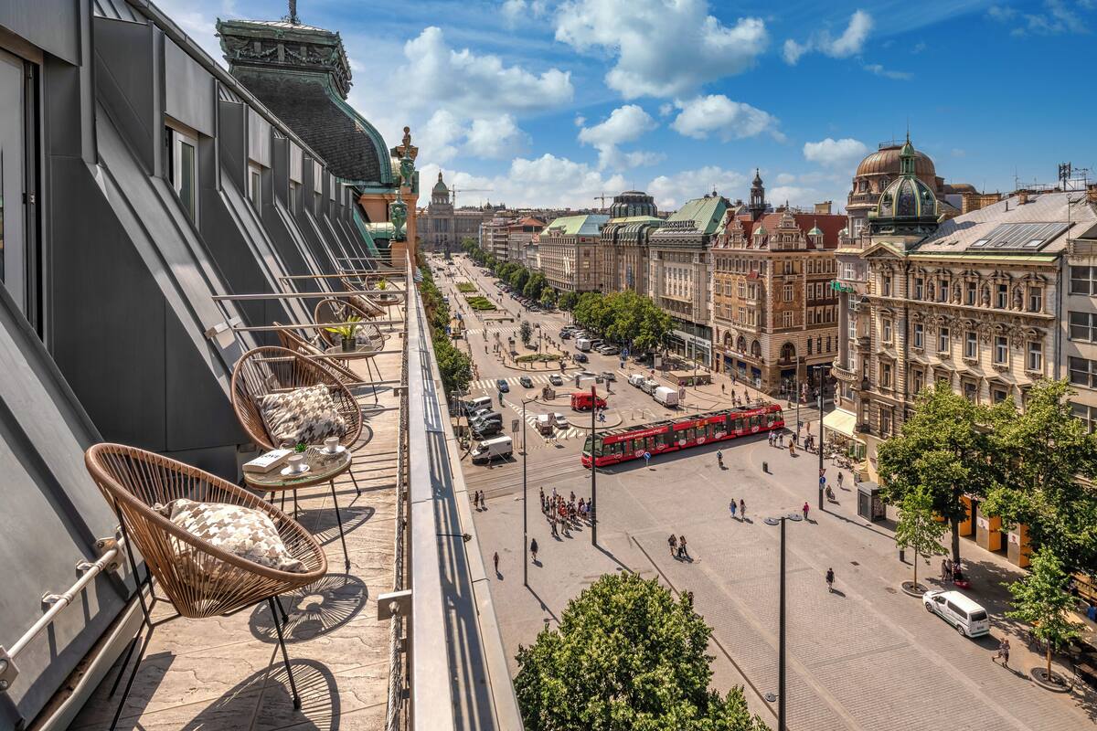 A balcony with wicker chairs and a table, overlooking a city street with historic buildings and a red tram.