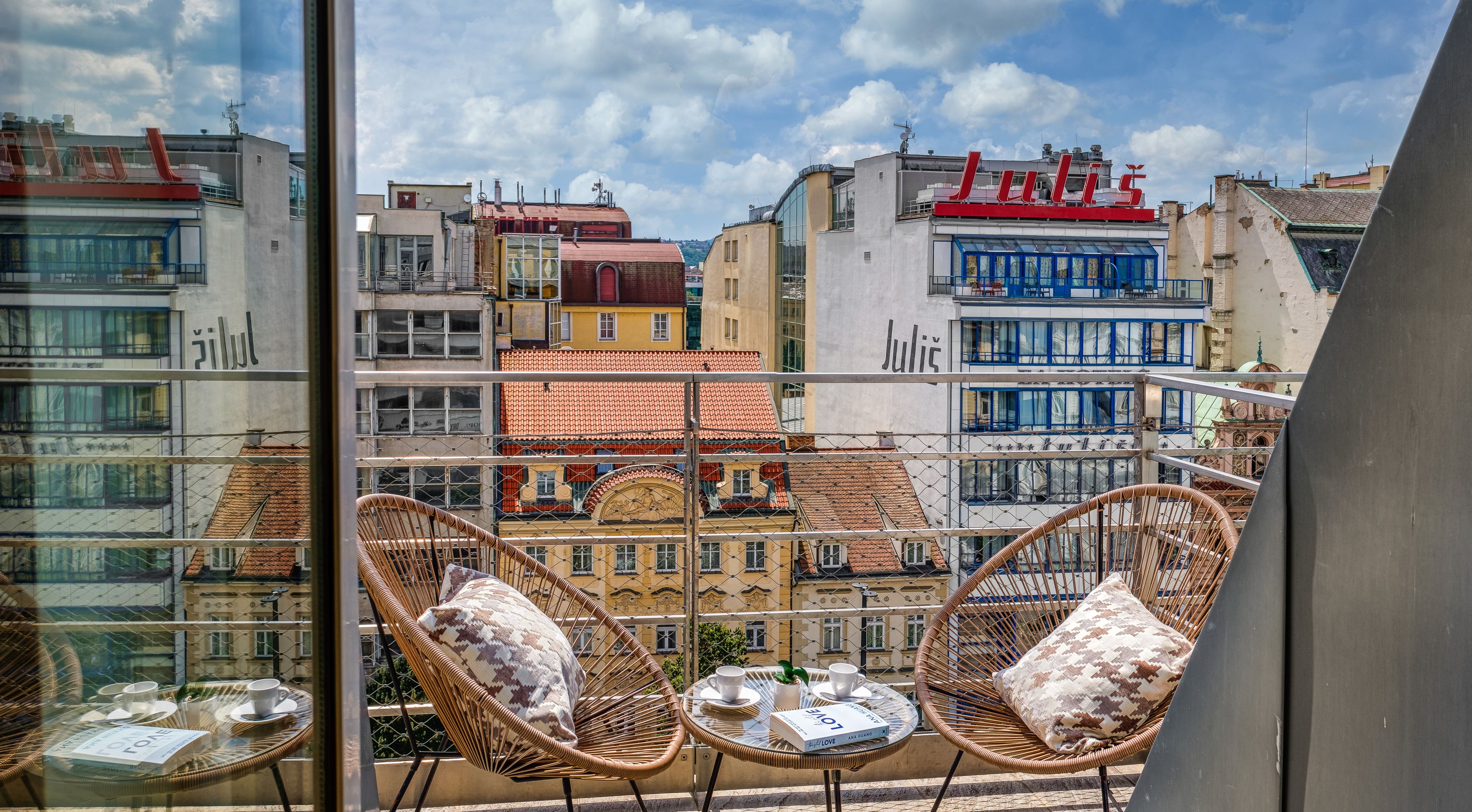 Un balcón con sillas de mimbre y una mesa, con vista a un paisaje urbano con edificios identificados como 'Julius' y 'Jülich'.