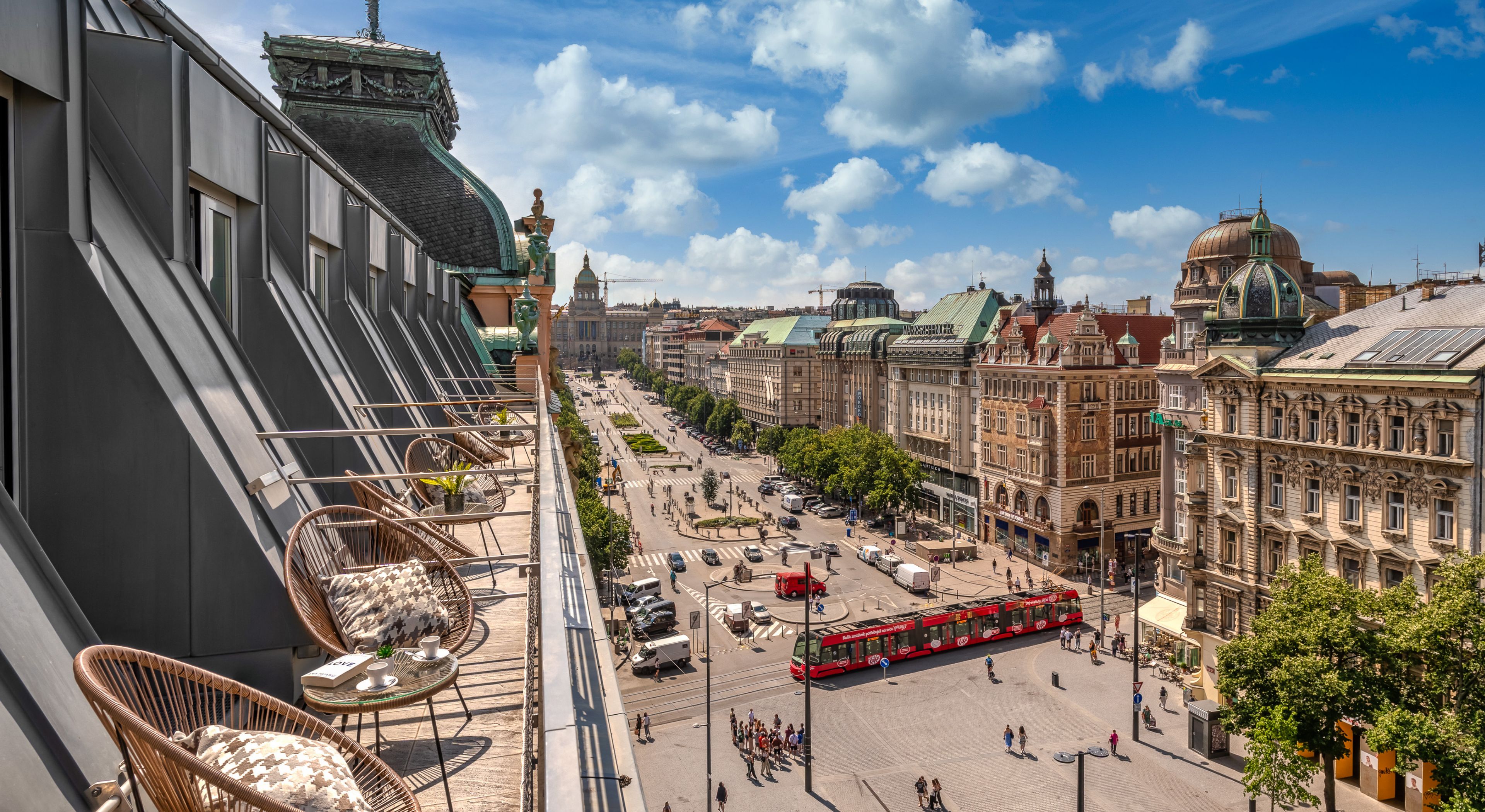 A view from a balcony overlooking a city square with historic buildings, a red tram, and a clear sky.