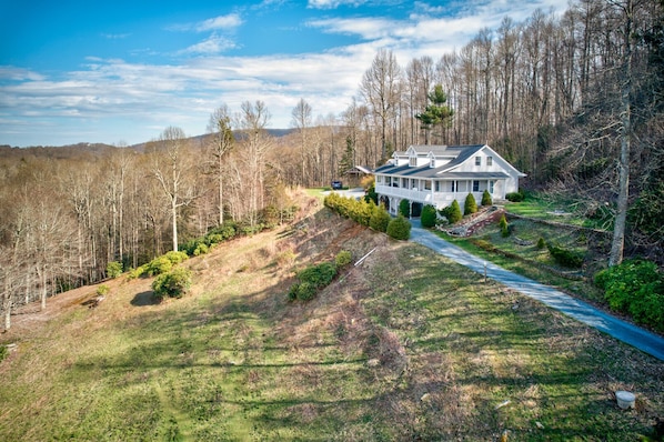 A large, single-story house with a wrap-around porch, surrounded by a forested hillside and a clear sky.