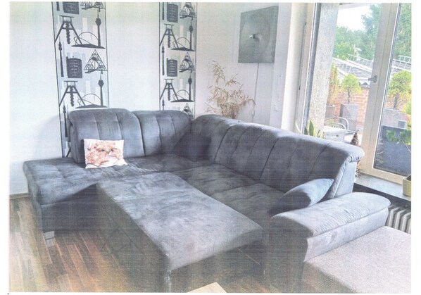 En grå hjørnesofa med et blomstret pudebetræk, en vægmonteret højttaler, og udsigt til en bro og grøntområde udenfor.