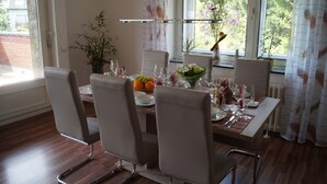 Uma área de refeições com uma mesa posta para quatro, cadeiras, um vaso com flores e uma janela com cortinas.