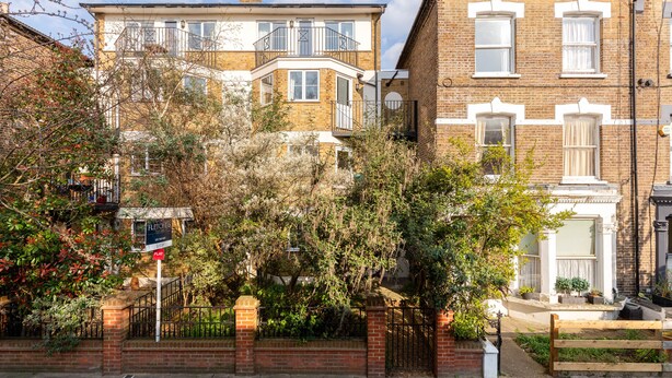A two-story residential building with a flat sign, a brick facade, and a small garden area with a wooden gate.