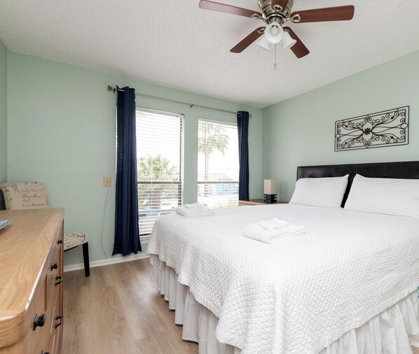 A hotel room with a large bed, a wooden dresser, a ceiling fan, and a window with curtains.