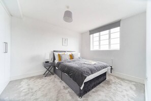A modern bedroom with a grey bed, white walls, a window, and a ceiling light. - Livestay- Perfect For Contractors Luxury Apartment (Slough)